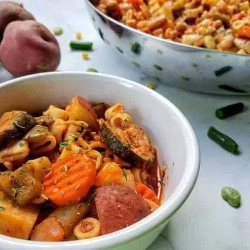 Vegetable ditalini soup served in a bowl. Bowl sits next to some of the raw ingredients and a large pan of the soup, all on a white marble background.