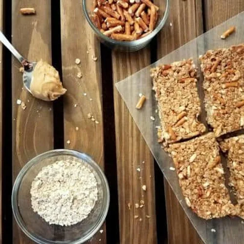 No-bake peanut butter oatmeal granola bars sit on a piece of wax paper on a dark wood plank background. Adjacent to the bars is a spoon with a scoop of peanut butter, a small glass bowl of pretzels, and a small glass bowl of oats. Oats and pretzel pieces are sprinkled about.