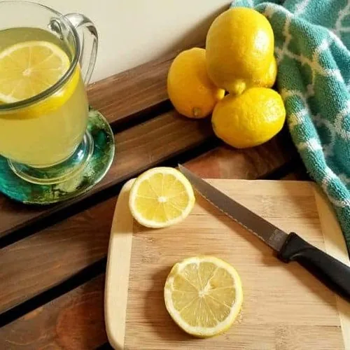 Lemon honey tea in a glass mug sitting on a coaster. On a small wooden cutting board rests a serrated knife and some lemon slices. In the background is a pile of lemons and a turquoise colored towel.