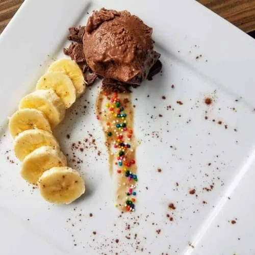 Vegan chocolate peanut butter banana ice cream on a square white plate. The ice cream rests on a bed of homemade chocolate chips and is flanked on one side by banana slices. Cocoa powder is sprinkled over the entire plate.