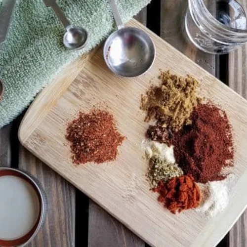 Piles of individual spices that make up homemade taco seasoning sit on a small cutting board on a dark wood plank background. Off to the side are measuring spoons and a small mason jar.