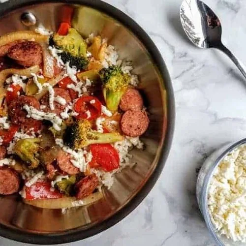 Vegetables, rice, and kielbasa in a stainless steel skillet sitting next to a small glass bowl of shredded mozzarella and a single stainless steel spoon. Everything sits on a white marble background.