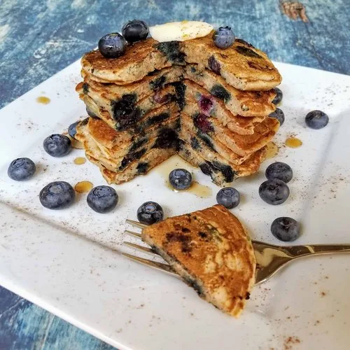 A stack of blueberry buttermilk pancakes on a square white plate sitting on a blue background. The pancakes are topped with a pat of homemade butter and are lightly drizzled with maple syrup.