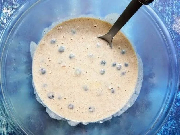Blueberry Buttermilk Pancakes Process Step 3c Blueberries uniformly distributed in pancake batter in a large glass bowl on a blue background.