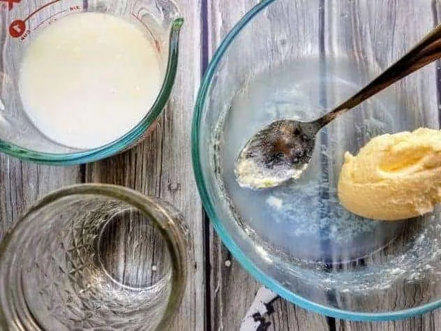 Buttermilk in a Pyrex measuring cup, homemade butter in a small glass bowl, and cold water in a small mason jar. All three sit on a gray wood plank background.