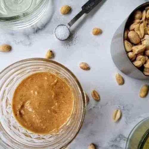 Homemade peanut butter in a small mason jar on a white marble background. A cup of peanuts, extra light olive oil in a small glass dish, maple syrup in a small glass dish, and a 1/4 teaspoon of Kosher salt sit nearby. Peanuts are also sprinkled about.