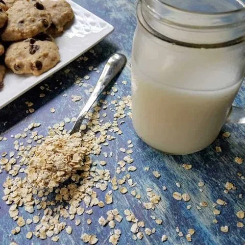 A mason jar filled with homemade oat milk sits on a blue background. A white plate of chocolate chip oatmeal pulp cookies sits in the upper left corner, and a spoon overflowing with uncooked oats sits next to the mason jar.