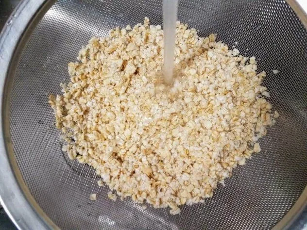 Soaked oats being rinsed under running water in a fine mesh stainless steel sieve.