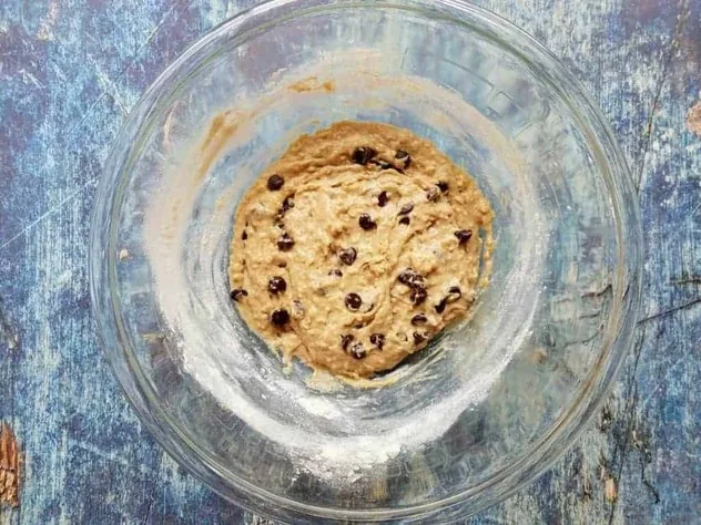 Chocolate Chip Oatmeal Pulp Cookies Process Step 3b Addition of chocolate chips to the mixture in a large glass bowl sitting on a blue background.