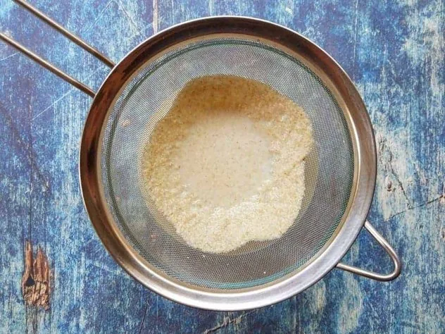 Straining oat milk through a fine mesh stainless steel sieve over a large glass bowl on a blue background.