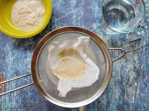 Straining oat milk through a cheesecloth covered fine mesh stainless steel sieve over a large glass bowl on a blue background. Oat pulp sits in a green bowl in the upper left corner and 1 cup of water sits in a Pyrex measuring cup in the upper right.