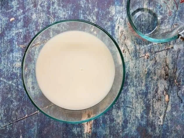 Finished homemade oat milk sits in a large glass bowl with an empty Pyrex measuring cup in the upper right corner. Everything sits on a blue background.