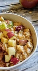 Microwave apple peanut butter oatmeal in a small white bowl on a wood background. Oatmeal is topped with vertical strips of chopped walnuts, cinnamon, and apples. To the right is a spoon with peanut butter. Apples sit in the background, with chopped walnuts and oatmeal sprinkled around.