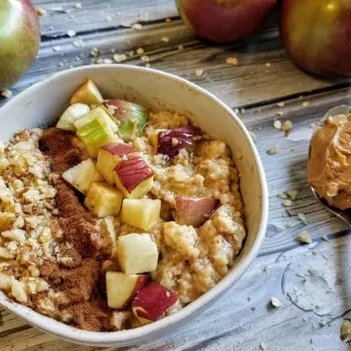 Microwave apple peanut butter oatmeal in a small white bowl on a wood background. Oatmeal is topped with vertical strips of chopped walnuts, cinnamon, and apples. To the right is a spoon with peanut butter. Apples sit in the background, with chopped walnuts and oatmeal sprinkled around.