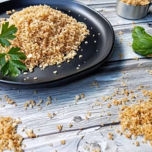Homemade shake and bake on a black plate on a wood plank background. In the foreground are small piles and crumbs of shake and bake. A measuring cup with shake and bake sits in the upper right corner. A sprig of parsley sits on the plate and a sprig of basil sits off to the right.