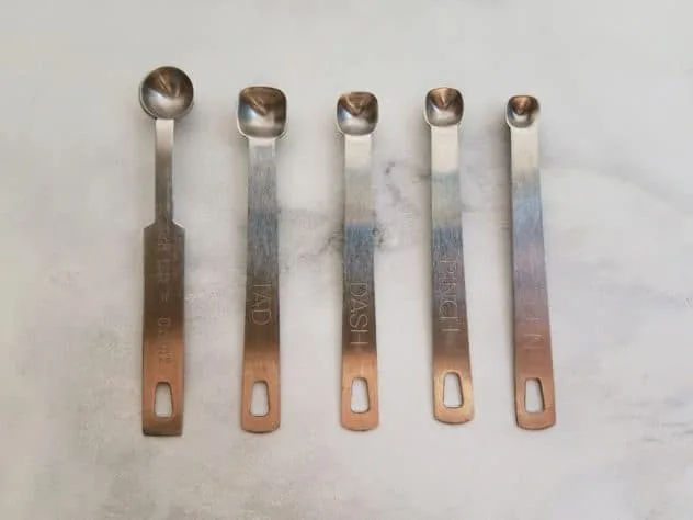 Side-by-side comparison of the 1/8, tad, dash, pinch, and smidgen measuring spoons. Spoons sit on a white marble background.