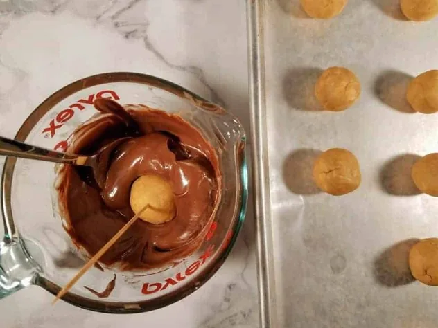 Chocolate Peanut Butter Balls Process Step 5a 3/4" uncoated peanut butter balls on a wax paper lined baking tray sit on the right. The glass Pyrex measuring cup with melted chocolate sits on the left. A peanut butter ball speared with a toothpick is being dipped and coated in the melted chocolate.