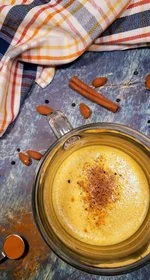 A glass of golden milk latte sits in the lower right. A dish towel with orange, yellow, and navy blue stripes sits in the upper left. Almonds, black peppercorns, and cinnamon sticks are strewn about. A 1/2 teaspoon of turmeric sits in the lower left.
