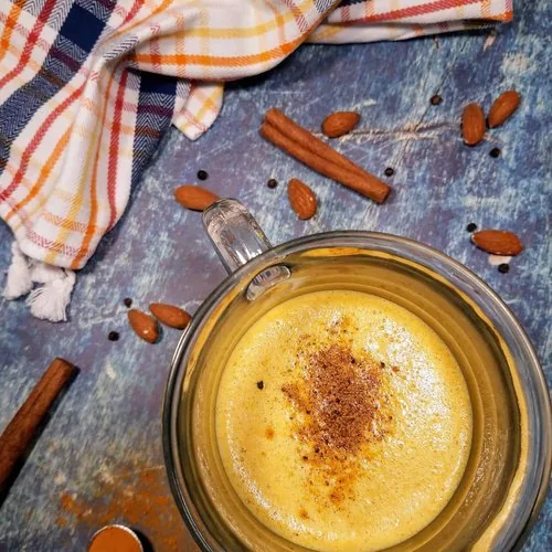 A glass of golden milk latte sits in the lower right. A dish towel with orange, yellow, and navy blue stripes sits in the upper left. Almonds, black peppercorns, and cinnamon sticks are strewn about. A 1/2 teaspoon of turmeric sits in the lower left.