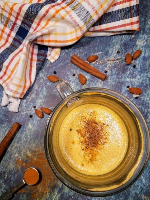 A glass of golden milk latte sits in the lower right. A dish towel with orange, yellow, and navy blue stripes sits in the upper left. Almonds, black peppercorns, and cinnamon sticks are strewn about. A 1/2 teaspoon of turmeric sits in the lower left.
