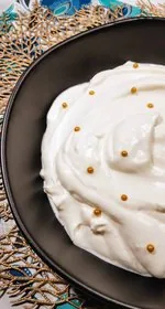 Homemade whipped cream sits in a large black bowl. The whipped cream is decorated with gold spherical sprinkles. The black bowl sits on top of a gold leaf placemat, which sits on top of a teal, navy, gray, black, and white colored paisley placemat, which sits on top of the white marble background. This is the vertical image.