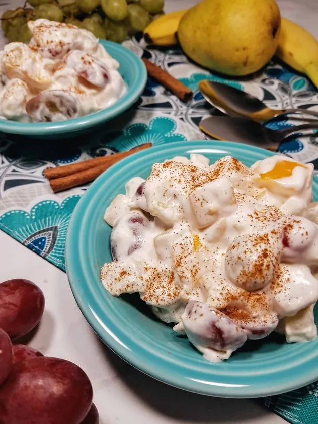 Whipped cream fruit salad topped with ground cinnamon sits in two teal colored fruit bowls. In the lower left is a handful of red grapes. In the background are two stainless steel spoons, a pear, a banana, and a bunch of green grapes. Two cinnamon sticks sit between the fruit bowls. Everything sits on a teal, navy, gray, black, and white colored paisley placemat which sits on top of a white marble background. This is the vertical image.