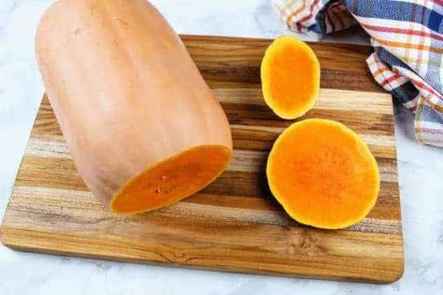 Baked Butternut Squash Process Step 1 A butternut squash with both of its ends cut off, sits on a wooden cutting board. A striped dish towel sits in the upper right corner. Everything sits on a white marble background.
