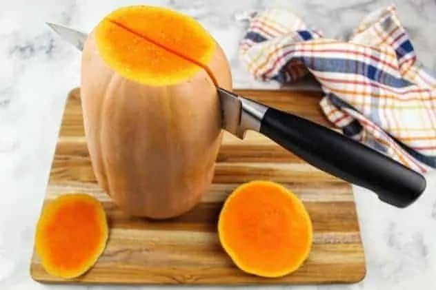 Baked Butternut Squash Process Step 2a A butternut squash stands up on its end on a wooden cutting board, with a chef's knife partway down it lengthwise. The chef's knife is centered on the butternut squash. A striped dish towel sits in the upper right corner. Everything sits on a white marble background.