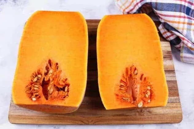 Baked Butternut Squash Process Step 2b A butternut squash halved lengthwise, sits on a wooden cutting board on a white marble background. A striped dish towel sits in the upper right corner.