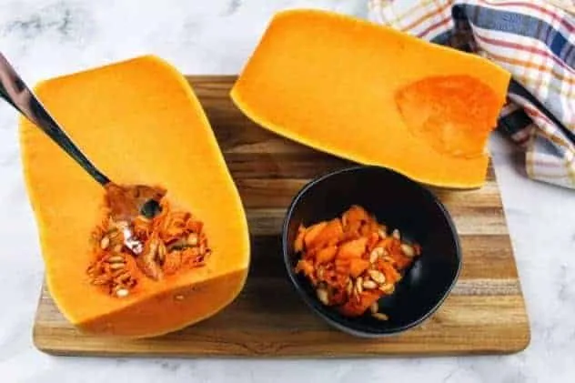 Baked Butternut Squash Process Step 3 The two butternut squash halves sit side-by-side, one with its seeds scooped out and the other is in the process of having its seeds scooped. A small black bowl contains the scooped seeds. Everything sits on a wooden cutting board. A striped dish towel sits in the upper right corner. Everything sits on a white marble background.
