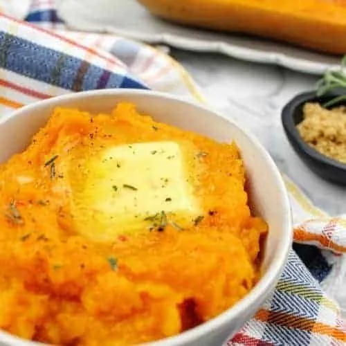 Mashed baked butternut squash topped with a pat of butter, rosemary, salt, and pepper, fills a small white bowl which sits on top of a striped dish towel. In the background is a tiny black bowl with light brown sugar and a sprig of rosemary. Also in the background is a white serving platter with half of a baked butternut squash. Everything sits on a white marble background. This is the horizontal image.