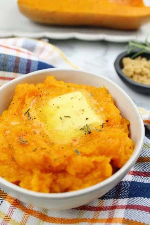 Mashed baked butternut squash topped with a pat of butter, rosemary, salt, and pepper, fills a small white bowl which sits on top of a striped dish towel. In the background is a tiny black bowl with light brown sugar and a sprig of rosemary. Also in the background is a white serving platter with half of a baked butternut squash. Everything sits on a white marble background. This is the vertical image.