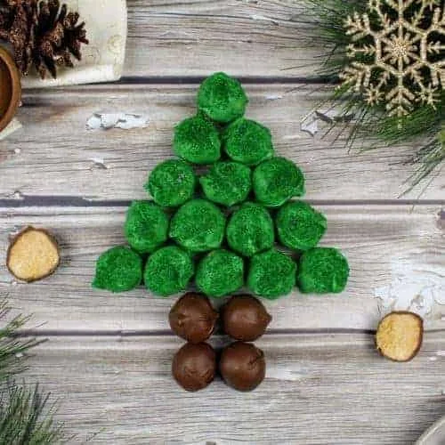 In the center are green and brown colored chocolate peanut butter balls arranged in the shape of a Christmas tree. Near the Christmas tree is an open peanut butter ball. Sprigs of fake evergreen trees sit in the lower left and the upper right. A gold snowflake also sits in the upper right. In the lower right are more peanut butter balls on a white plate. In the upper left is an ivory colored piece of cloth with gold glitter swirls, a cluster of pine cones, and a small dark brown wooden bowl of green sparkling sugar sprinkles. Everything sits on a wooden plank background. This is the horizontal image.