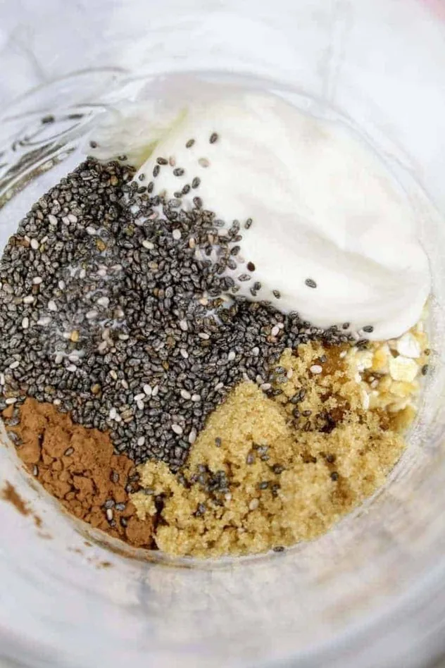 Chocolate Overnight Oats Process Step 2a Overhead view of chia seeds, Greek vanilla yogurt, quick oats, light brown sugar, and unsweetened cocoa powder sitting on top of the unsweetened almond milk in a glass mason jar. Everything sits on a white marble background.