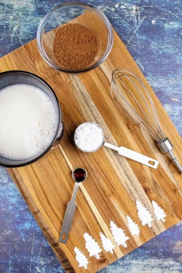 Starting from the left and moving clockwise: a navy blue mug filled with almond milk that has been warmed up, a small glass bowl with cocoa powder, a small whisk, a tablespoon of confectioner's sugar, and a 1/4 teaspoon of vanilla extract all sit on a wooden cutting board. At the bottom of the cutting board are pine trees made of confectioner's sugar. The cutting board sits on a navy blue background.