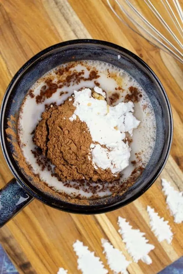 Addition of cocoa powder, confectioner's sugar, and vanilla extract to the mug filled with almond milk. The whisk sits in the upper right. At the bottom of the cutting board are pine trees made of confectioner's sugar. The cutting board sits on a navy blue background.