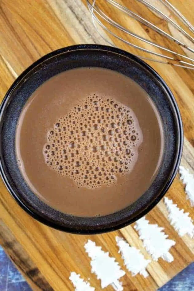 Vegan hot cocoa after mixing everything in and heating it up to the desired temperature. The whisk sits in the upper right. At the bottom of the cutting board are pine trees made of confectioner's sugar. The cutting board sits on a navy blue background.