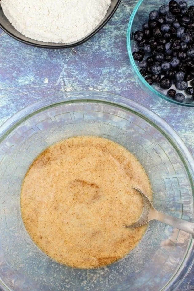 Vegan Blueberry Muffins Process Step 1 At the bottom is a large glass mixing bowl with baking powder, kosher salt, ground cinnamon, vegan butter, extra light olive oil, applesauce, and almond milk all mixed together. In the upper right is a small bowl of blueberries and in the upper left is a black bowl with gluten-free flour. Everything sits on a navy blue background.