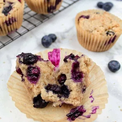 In the center of the picture is a blueberry muffin that has been split open to show the inside. The split halves sit in the paper baking cup. In the background is one other vegan blueberry muffin and a navy and white checkered tea towel in the upper right. In the upper left is a cooling rack with more muffins. Blueberries and demerara sugar are sprinkled about. Everything sits on a white marble background.