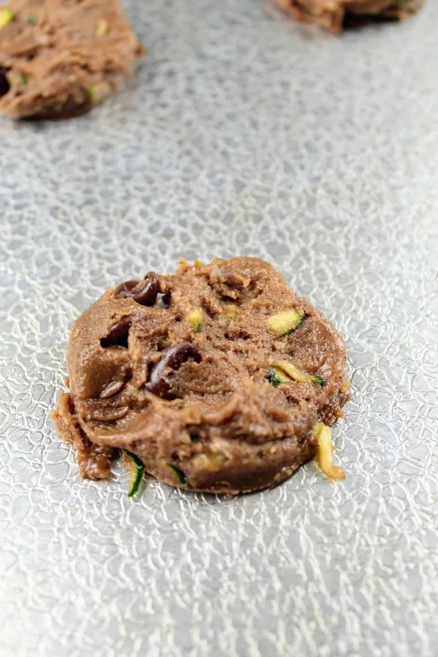 A slightly flattened vegan chocolate zucchini cookie on a textured aluminum baking sheet before baking.
