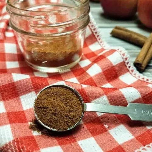 A teaspoon of apple pie spice sits in the foreground, with a jar of the spice, four apples, and two cinnamon sticks sit in the background. The jar and the teaspoon sit on a red and white checkered kitchen towel, whereas the apples and cinnamon sticks sit on a wood plank background. This is the vertical image.