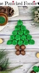 In the center are green and brown colored chocolate peanut butter balls arranged in the shape of a Christmas tree. Near the Christmas tree is an open peanut butter ball. Sprigs of fake evergreen trees sit in the lower left and the upper right. A gold snowflake also sits in the upper right. In the lower right are more peanut butter balls on a white plate. In the upper left is an ivory colored piece of cloth with gold glitter swirls, a cluster of pine cones, and a small dark brown wooden bowl of green sparkling sugar sprinkles. Everything sits on a wooden plank background. At the top of the image are the words 'The Panicked Foodie,' with the i's colored red and dotted with red hearts. At the end is a black mixing bowl with a red heart on it that contains three sugar cubes.