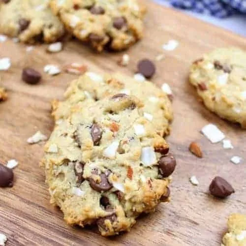 Seven vegan almond coconut chocolate chip cookies sit on a dark wooden serving board. Chocolate chips, chopped almonds, and chopped coconut are scattered about on the serving board. In the upper right is a navy blue and white checkered tea towel.