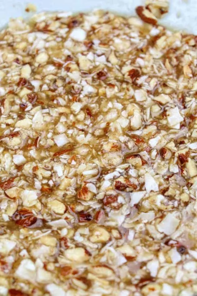 A mixture of extra light olive oil, granulated sugar, light brown sugar, unsweetened applesauce, vanilla extract, baking soda, Kosher salt, chopped coconut, and chopped almonds in a large glass bowl.