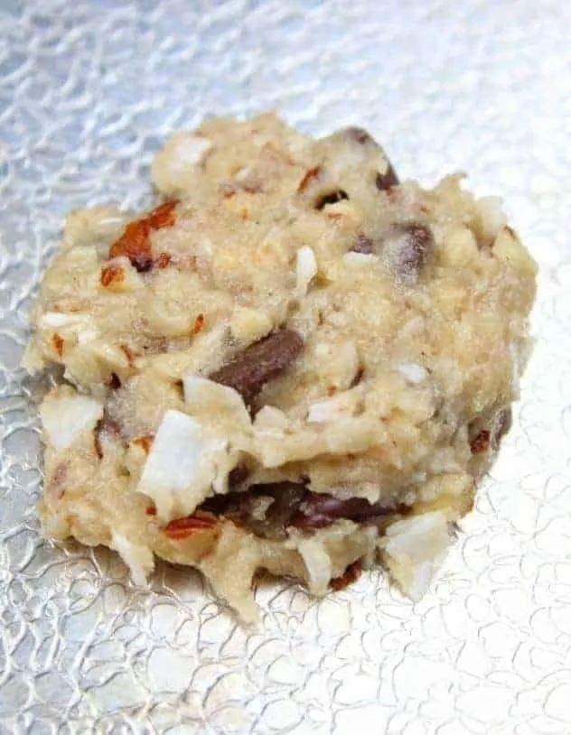 A single, flattened, unbaked vegan almond coconut chocolate chip cookie on a textured aluminum baking sheet.
