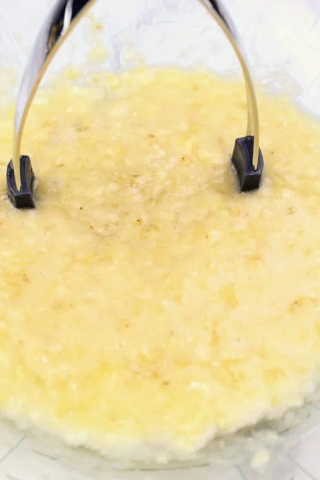 Mashed bananas and a potato masher sit in a large glass bowl.