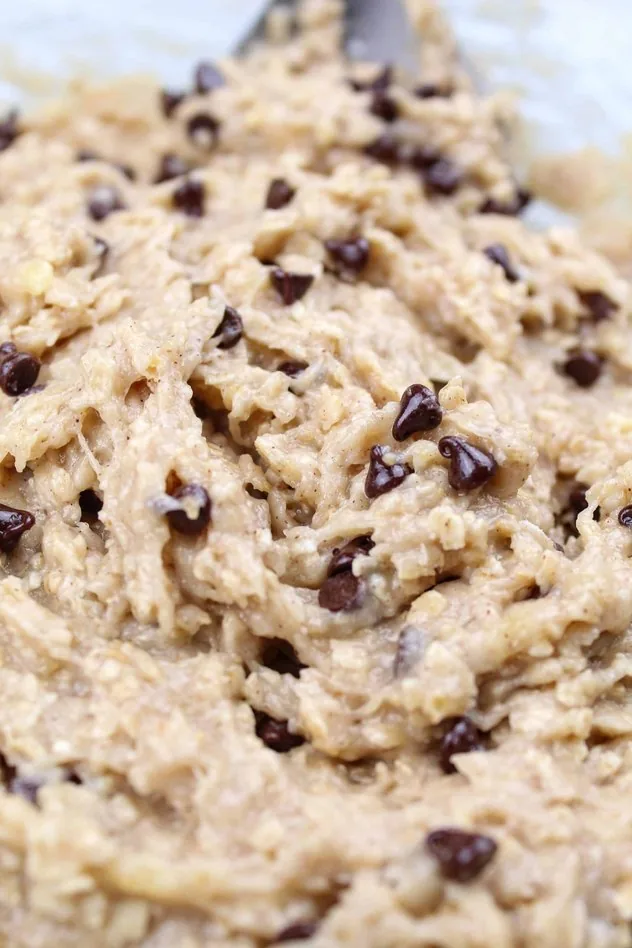 Addition of mini chocolate chips to the dough in a large glass mixing bowl with a stainless steel mixing spoon in the upper right.