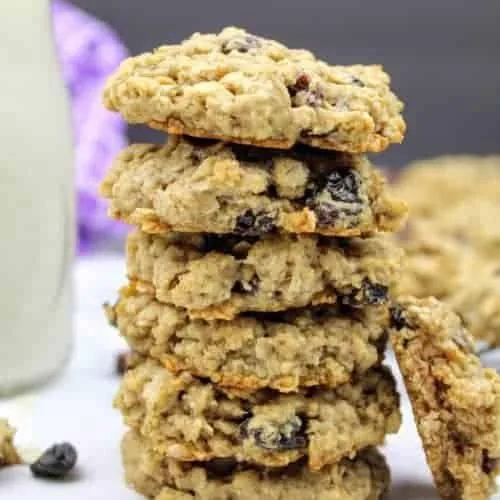 A stack of six vegan oatmeal raisin cookies sits in the center of the photo. Leaning against the stack on the right hand side is a half of a cookie to show the chewy interior. To the left of the stack, is a small glass of almond milk. In the background on the left is a purple tea towel and on the right are more cookies sitting on a cooling rack. Everything sits on a white marble background with a black vertical background. Scattered about are oat grains and raisins.