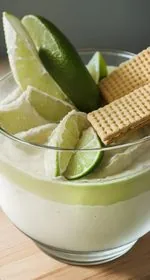 In the center is a small black bowl containing vegan key lime hummus garnished with coconut cream, lime zest, and crushed graham crackers. On the left side of the image are graham crackers and a lime in the lower left. In the upper right are some limes. In the lower right is a black plate of graham crackers. Sprinkled about are graham cracker crumbs and threads of lime zest. Everything sits on a white marble background. This is a vertical image taken from an overhead view. At the top of the image are the words 'The Panicked Foodie,' with the i's colored red and dotted with red hearts. At the end is a black mixing bowl with a red heart on it that contains three sugar cubes.