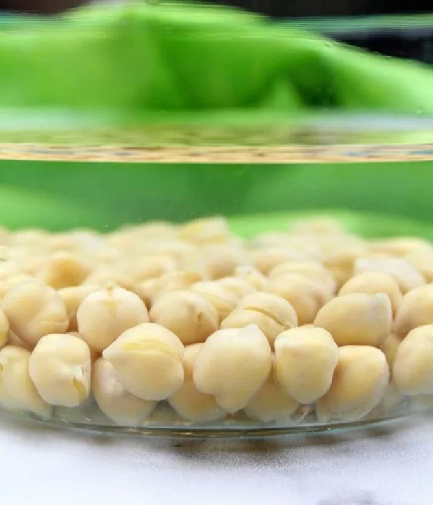 Chickpeas soak in a glass bowl filled with water. In the background is a lime green tea towel.